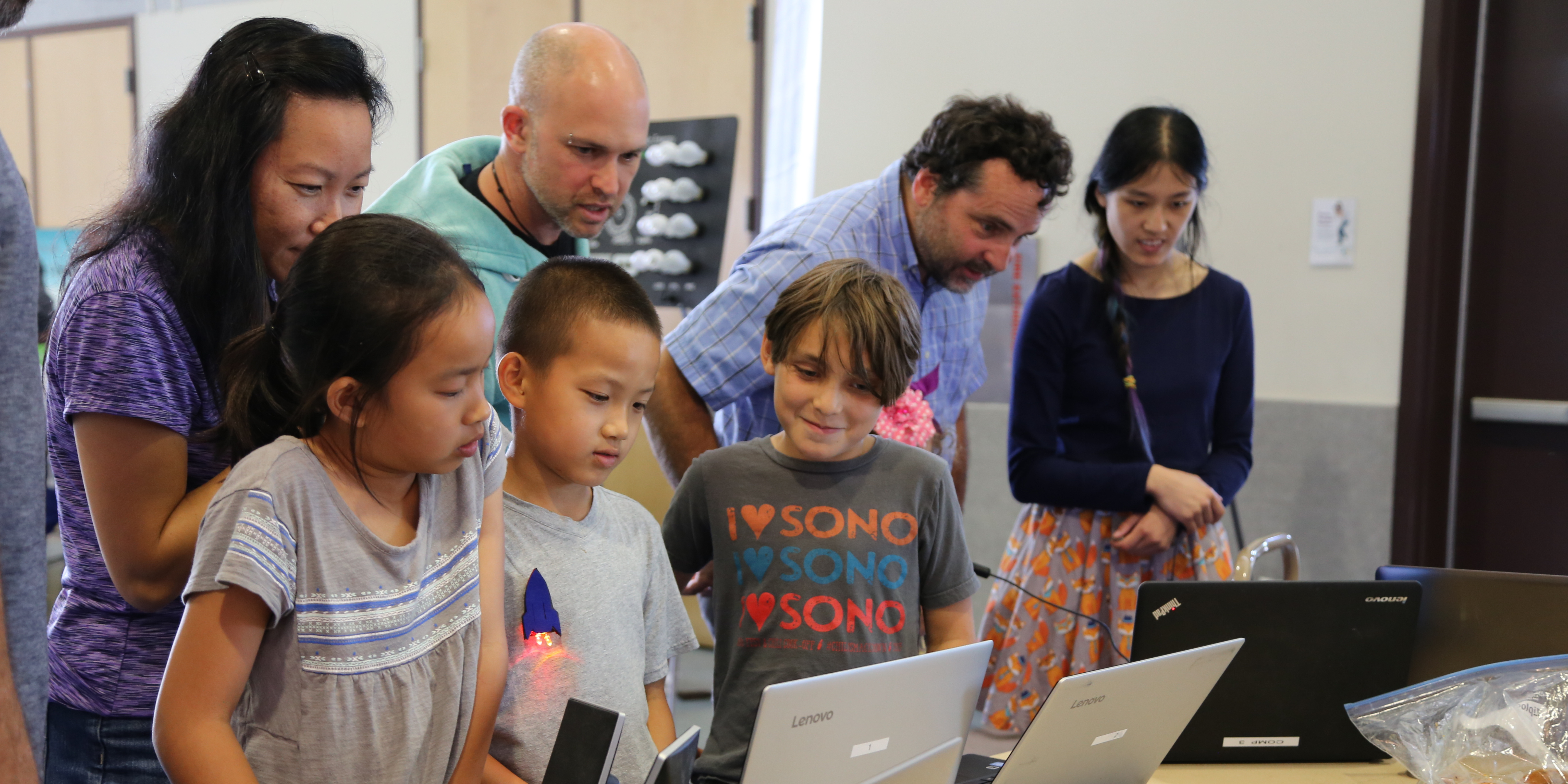 Children and adults surrounded around a computer.