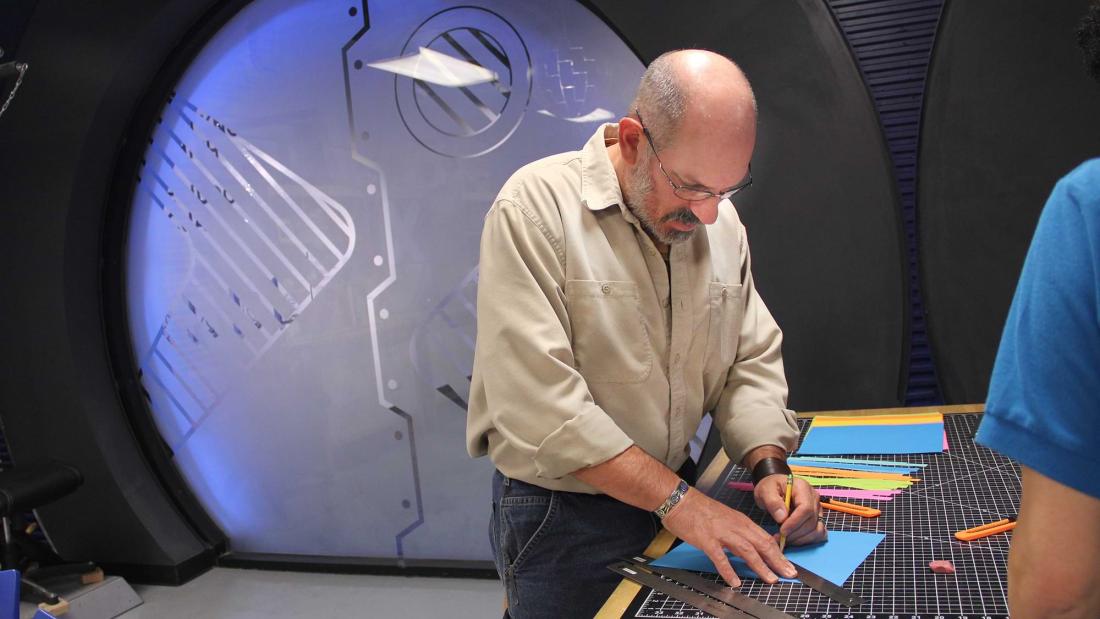 An adult man works on a wooden table with a cutting mat and multiple brightly colored construction paper pieces at the Fleet Science Center