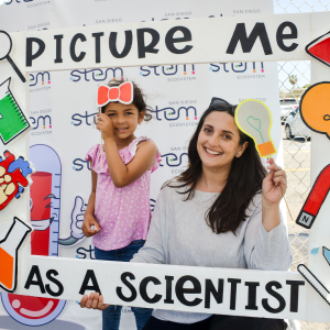 A woman and young girl pose behind a white frame that says 'picture me as a scientist'